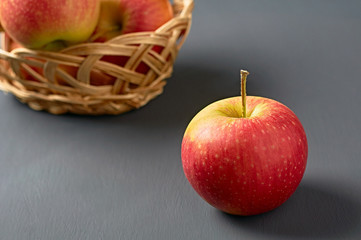 One red fresh whole ripe apple with vitamins on background of full wooden wicker basket and old dark scratched concrete table on kitchen. Harvest or diet concept. Close-up