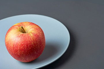 Single red fresh whole ripe apple full of vitamins lies on circle ceramic plate and old dark scratched concrete table on kitchen. Harvest or diet concept. Space for text