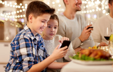 technology, holidays and people concept - happy boy with sister using smartphone having family dinner party at home