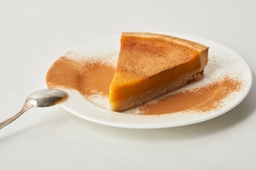 tasty pumpkin pie with cinnamon powder on plate near spoon of white marble table