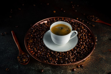 Round copper tray with kopi luwak coffee beans, wooden spoons, white cup of coffee with saucer on dark background