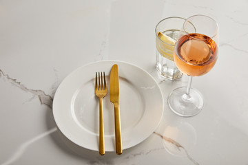 white plate with golden knife and fork near glasses with lemon water and rose wine on marble table