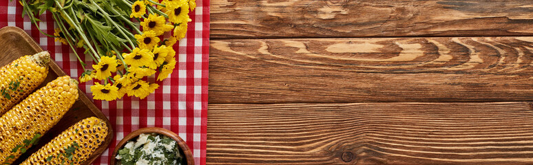 top view of grilled corn and yellow wildflowers on red checkered napkin on wooden table, panoramic shot