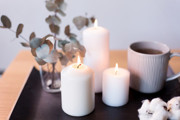Fototapeta na wymiar decoration, hygge and cosiness concept - burning white candles, tea in mug, branches of eucalyptus populus and cotton flowers on table