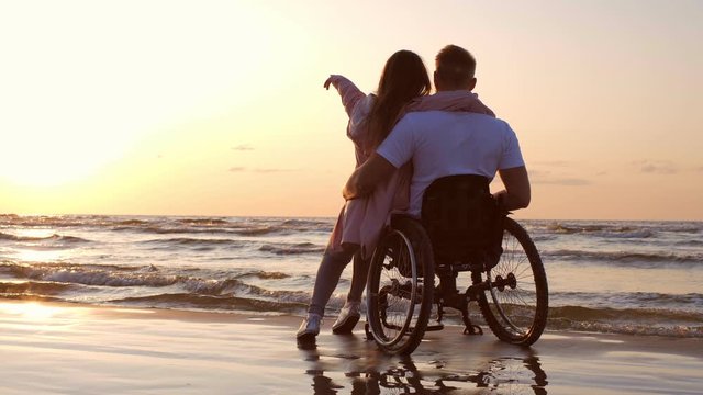 Handicapped man in wheelchair and his girlfriend on a beach at sunset