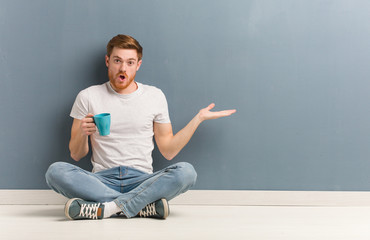 Young redhead student man sitting on the floor holding something on palm hand. He is holding a coffee mug.