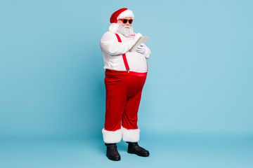 Full length photo of funny funky fat santa claus in hipster eyeglasses with big belly write wish list plan in note book use pen wear suspenders red pants boots isolated over blue color background