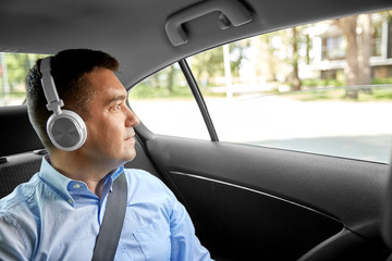transport, technology and people concept - male passenger or businessman with wireless headphones listening to music on back seat of taxi car