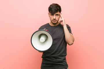 Young hispanic man holding a megaphone pointing his temple with finger, thinking, focused on a task.