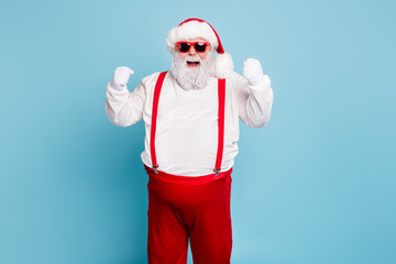 Fototapeta na wymiar Portrait of funny funky fat santa claus with big belly raise fists scream yeah have luck celebrate newyear x-mas party wear suspenders modern red spectacles isolated over blue color background