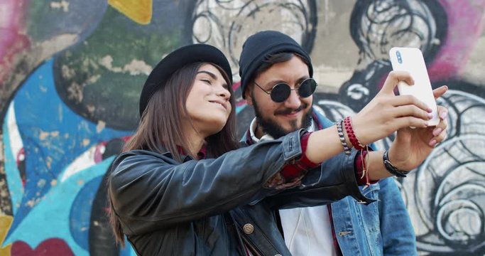 Portrait of the young attractive Caucasian hipsters couple taking a selfie photo with a smartphone camera at the graffity wall outdoor.