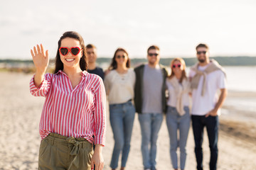 friendship, valentine's day and people concept - happy woman in heart-shaped sunglasses with group of friends on beach in summer waving hand
