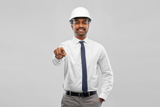 architecture, construction business and people concept - smiling indian male architect in helmet pointing finger to something over grey background