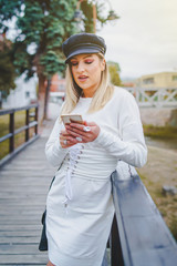 Young blond happy woman in white dress and black cap hat standing on the bridge in town or city with mobile phone using smart application or message in autumn or spring day