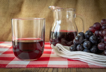 bicchiere con vino rosso, bricco e grappolo d'uva nera su tavolo con tovaglia a scacchi rossi e bianchi