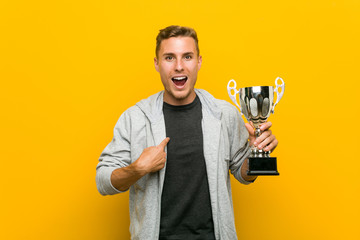 Young caucasian man holding a trophy surprised pointing at himself, smiling broadly.