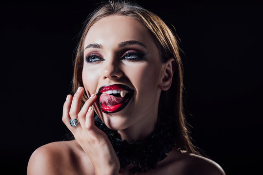 naked scary vampire girl with fangs licking fingers isolated on black