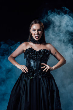 scary vampire girl with fangs in black gothic dress on black background with smoke