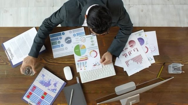 Top view shot of businessman in suit and glasses sitting at table and inspecting data on financial documents with graphs and charts, then typing on computer