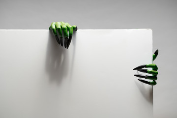 Two green zombie hands with long nails holds blank sheet of paper on gray background in studio for...
