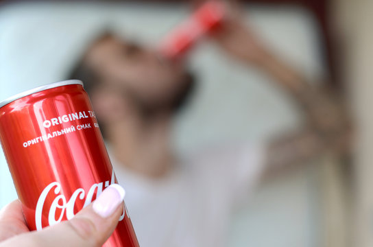 Caucasian Man Drinks Coca-Cola Drink In Garage Interior And Male Hand Presents One Coca Cola Red Can In Focus