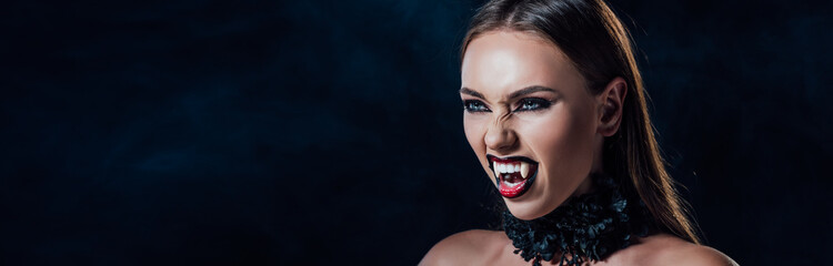 panoramic shot of scary vampire girl with fangs in black gothic dress isolated on black - obrazy, fototapety, plakaty