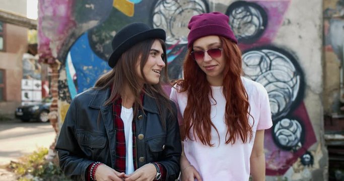 Two Caucasian young pretty and cheerful girls in hipsters style, best friends talking walking at the slums with graffity on walls.