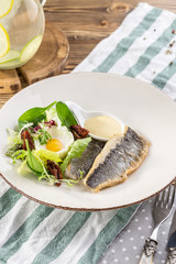 Fried white fish fillet with fried egg and greens on white plate on wooden table