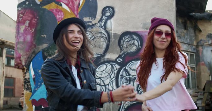 Best friends stylish hipsters walking the slums on the graffity wall background, friendly pretty girls talking, gossiping and laughing.