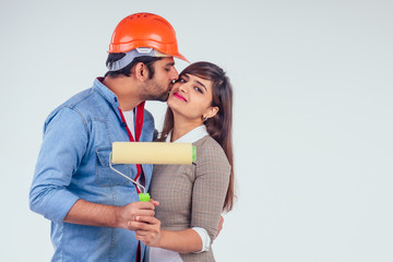 indian couple painting the walls with roll at their home white color background and wearing hardhat...