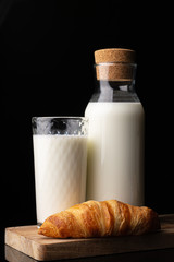 A glass with milk, a bottle with milk and one french croissant on a wooden board. Black background. Glass bottle. Natural cork. White drink. Cow's milk. Classic french croissant. Homemade baking.
