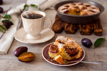 A piece of plum pie on a plate, tea in a cup, plums on a light timbered background.