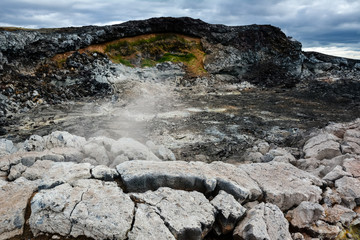 Leirhnjukur old black lava field with stones and smoke coming from ground and blue sky in Iceland,...