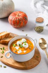 Pumpkin soup with seeds, mozzarella balls and greenery in white bowl on white background with autumn leaves and pumpkins. Close up. Menu, restaurant. Vegeterian seasonal food.