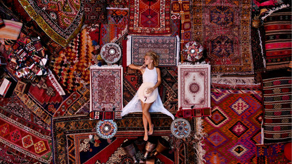 Beautiful happy girl in a long white dress laying on the carpet and rugs in Goreme, Cappadocia,...
