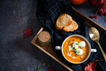 Pumpkin soup with seeds, mozzarella balls and greenery in white bowl on dark background with autumn leaves and pumpkins. Copy space, top view. Menu, restaurant. Vegeterian seasonal food.