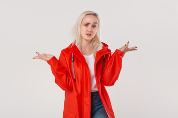 Blond girl in a red raincoat with hood expressing emotion of indignation  because of rainy weather isolated over white background. Get ready for bad weather.