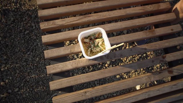 Top view of a female hand sticks chopsticks into noodles, a white take-out box with pasta and chicken is standing on a wooden deck chair on a pebble beach