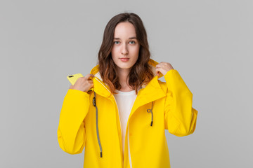 Smiling brunette girl gets on hood of a yellow raincoat isolated over white background. It is...