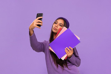 Pretty african-american teenager student in a hat and sweater holding folders and making selfie...