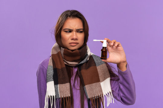 Pretty african-american girl in a sweater and cosy checked scarf got tired of using nose spray beacuase of cold standing isolated over purple background. Catching cold in winter.