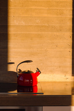 A red tea pot sits on a stove in front of the concrete wall of a kitchen at sunrise.