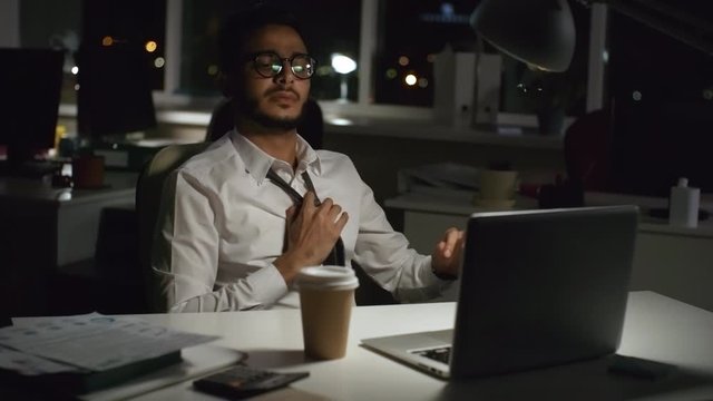 Medium shot of stressed Arab businessman in glasses typing on laptop in dark office while working overtime. He is exhaling in frustration and loosening his tie