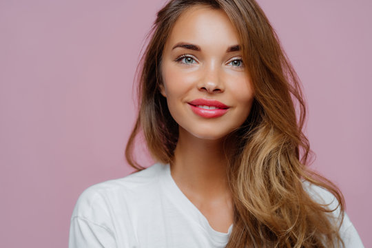 Horizontal cropped image of pleasant looking female model has tender smile, wears minimal makeup, has long wavy hair, looks at camera with satisfaction, models over violet wall, copy space for promo