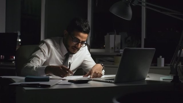 Medium shot of stressed Arab businessman in glasses sitting at his desk in dark office and writing ideas on sheet of paper, then crumpling it and throwing it into trash can in frustration