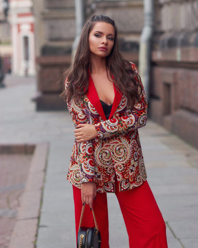 Body length outdoor portrait of young beautiful caucasian woman with long wavy brunette hair in red coat and trousers and sunglasses with black leather handbag posing at old city street