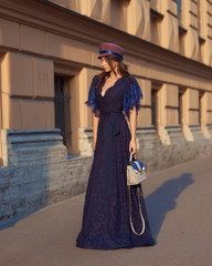 Fototapeta na wymiar Full length outdoor portrait of young beautiful elegant woman in long blue evening dress and purple hat with gray leather handbag standing and posing at old city street on a sunny evening day