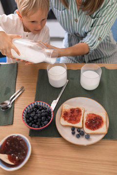 Cute boy and his mom pouring milk in his glass for breakfast at home
