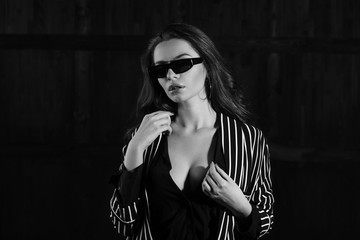 Female beauty concept. Closeup portrait of fashionable stylish young woman wearing black blouse, striped blazer and sunglasses posing over dark background. Perfect hair and skin. Black and white