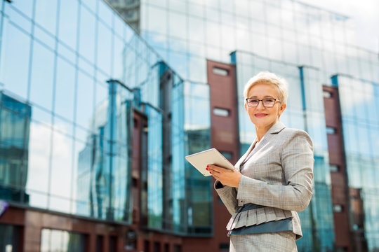 stylish adult woman in age in a suit and glasses against the background of a glass building with a tablet in her hands